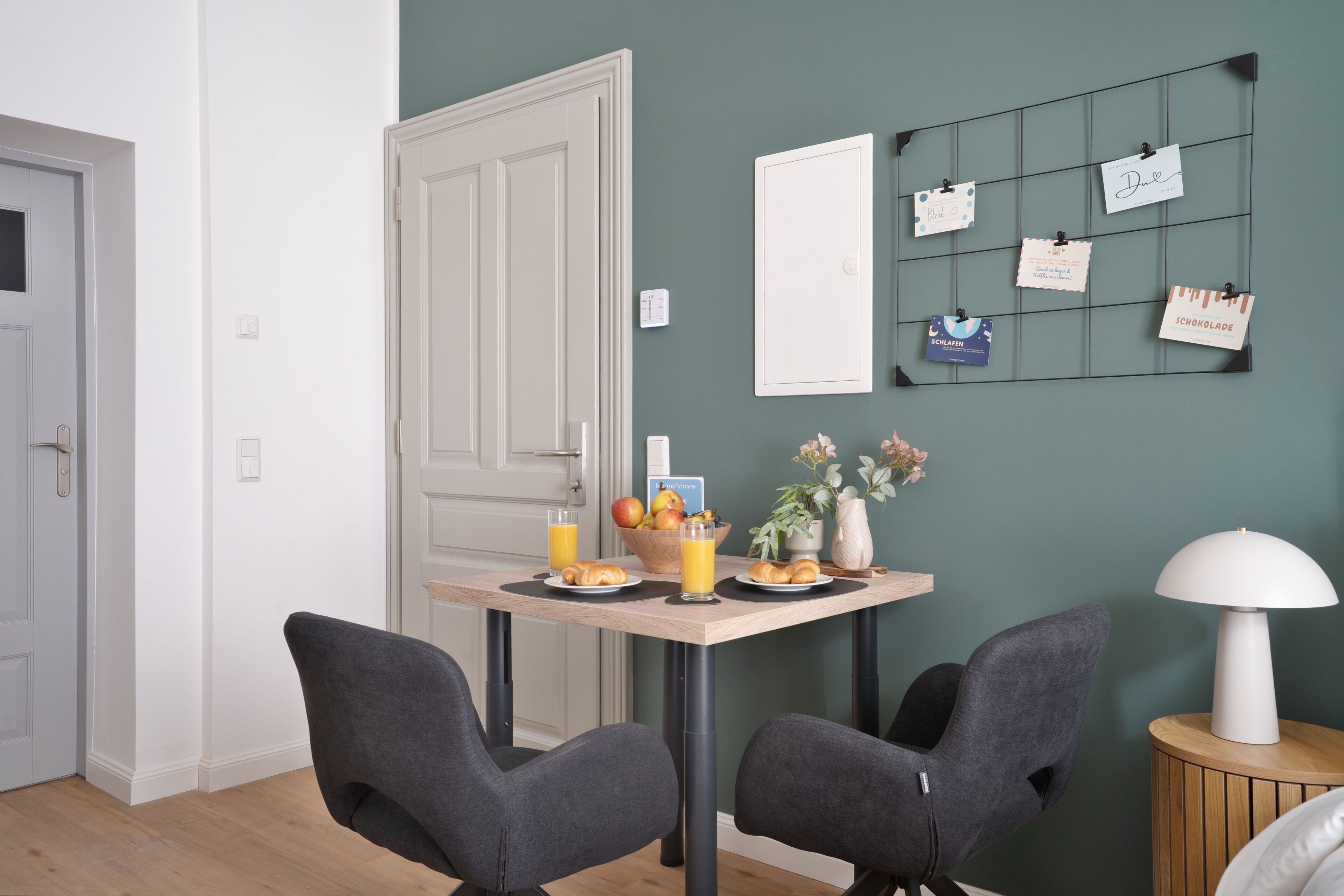 A dining area with a table set for two, a wall-mounted bulletin board with notes, and a lamp on a side table.