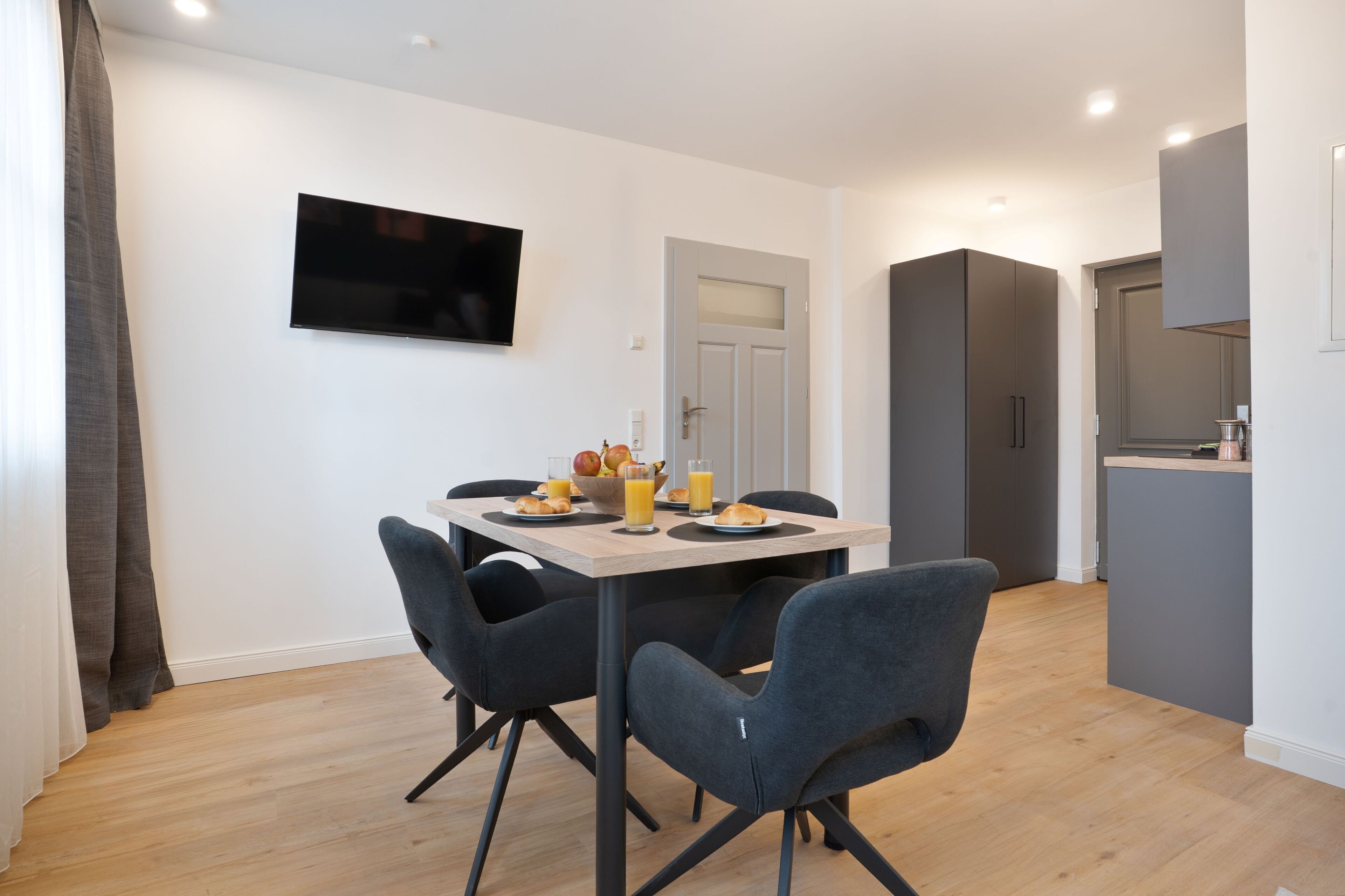 A modern dining area with a table set for four, a flat-screen TV mounted on the wall, and a kitchen area with a grey cabinet and countertop.