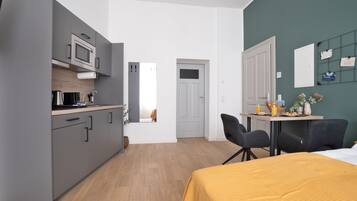 Une pièce compacte comprenant un coin cuisine, un lit recouvert d’une housse de couette jaune, une table à manger avec des chaises et un mur d’accent vert.