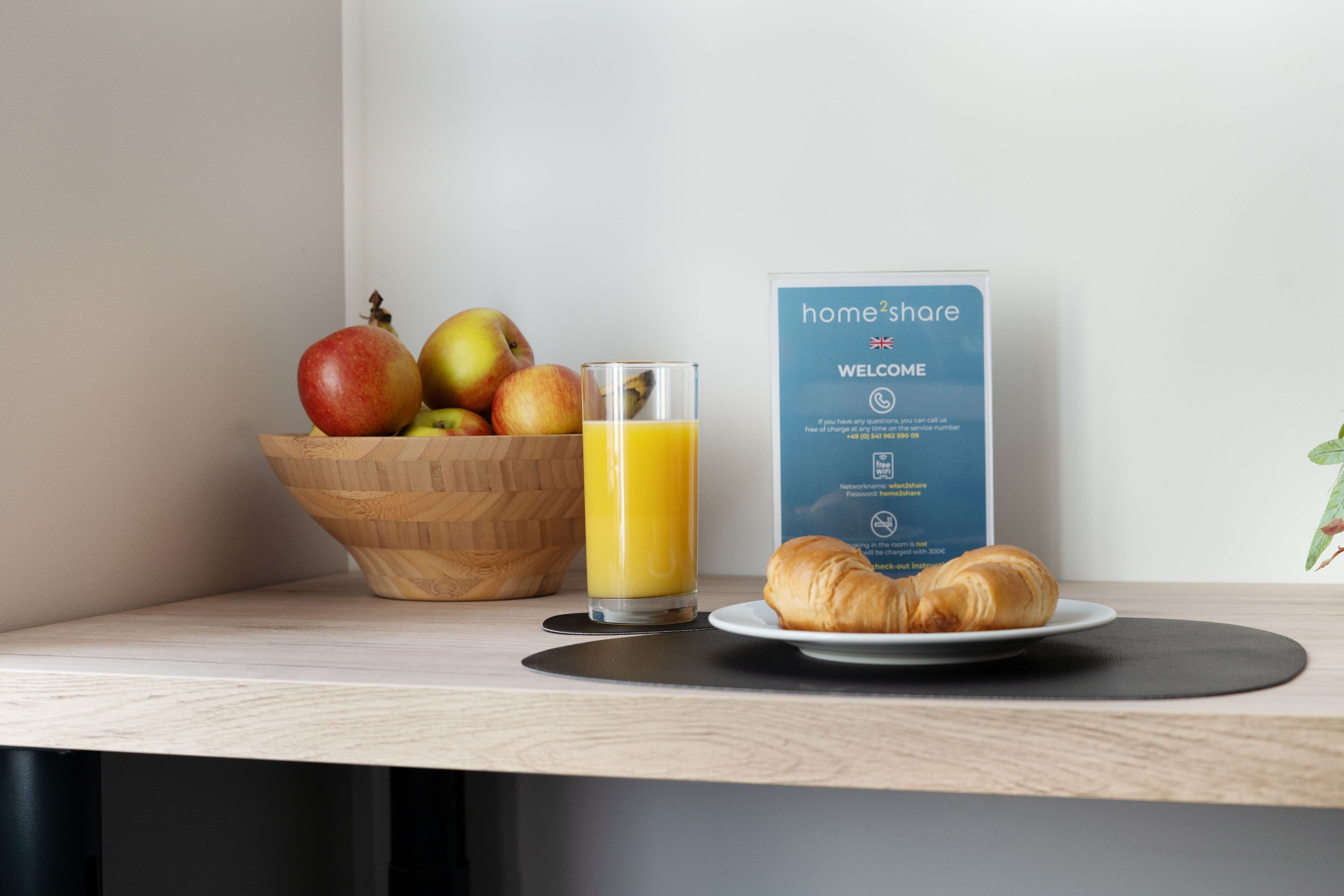 A breakfast setting with a wooden bowl of fruit, a glass of orange juice, and a croissant on a plate, alongside a welcome sign.