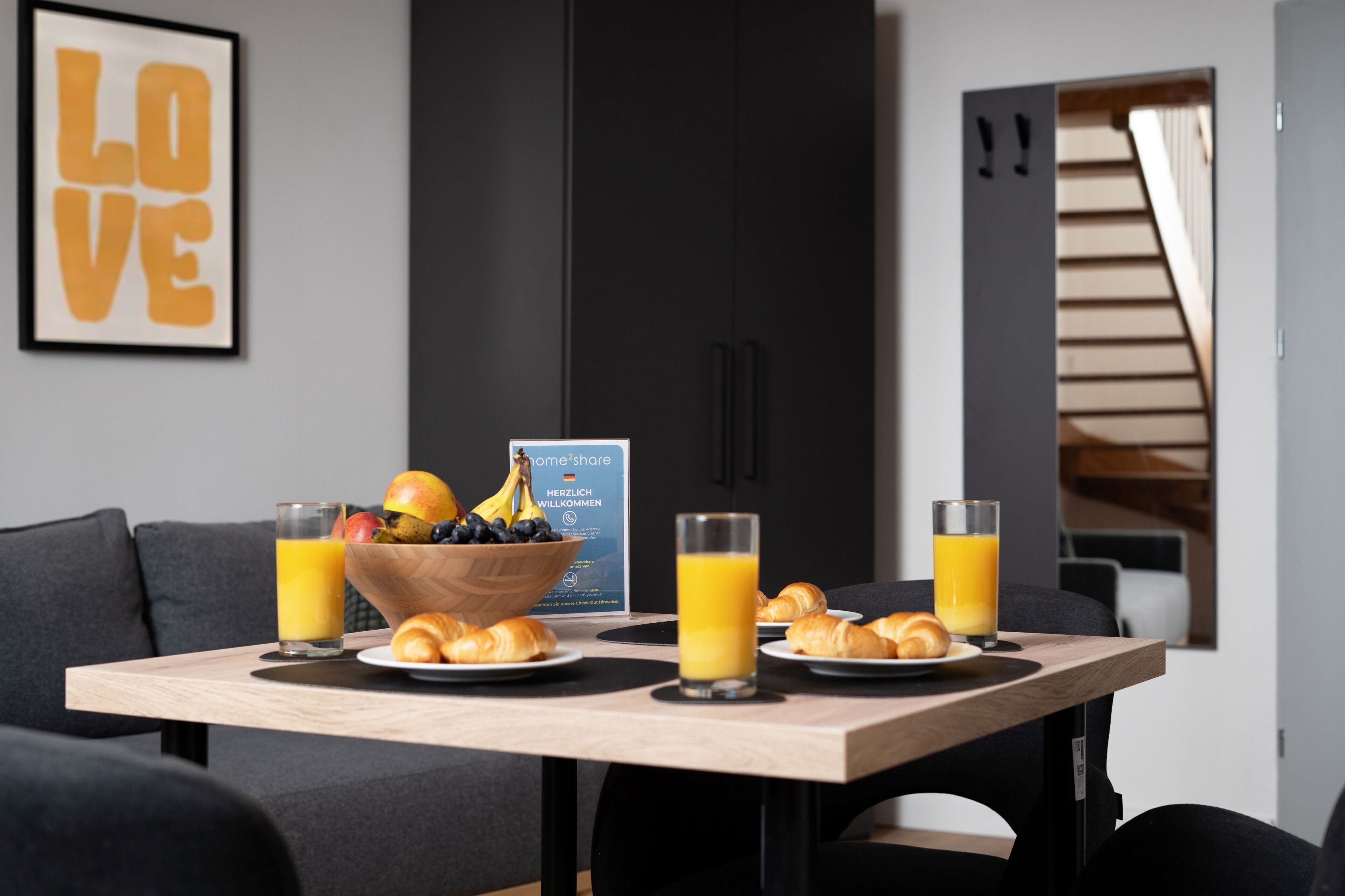 A breakfast setup with croissants, fruit, and orange juice on a table, with a staircase and a 'LOVE' sign in the background.
