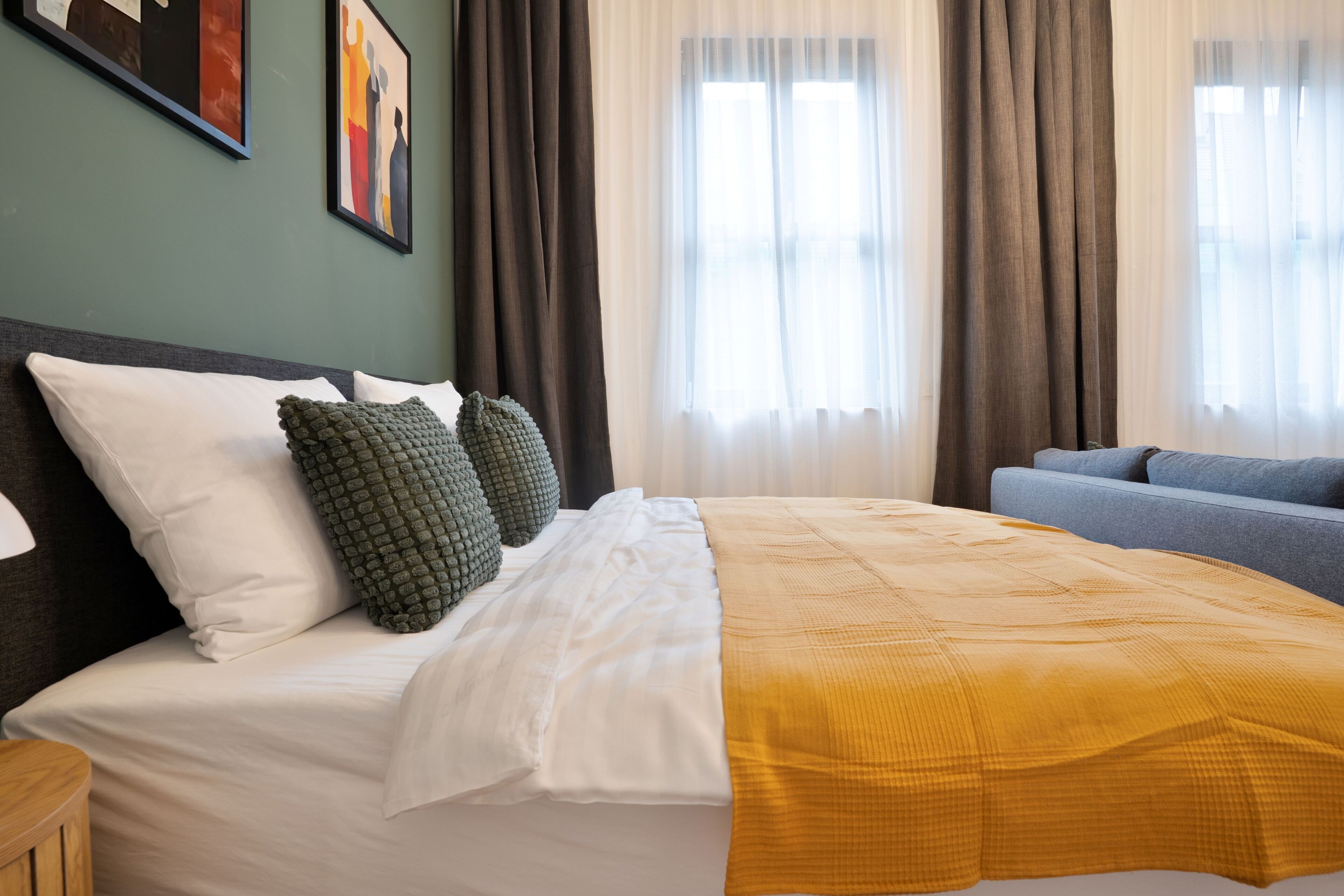 Ein Hotelzimmer mit einem Bett, einem grauen Sofa, einem Fenster mit Vorhängen und zwei gerahmten Kunstwerken an der Wand.