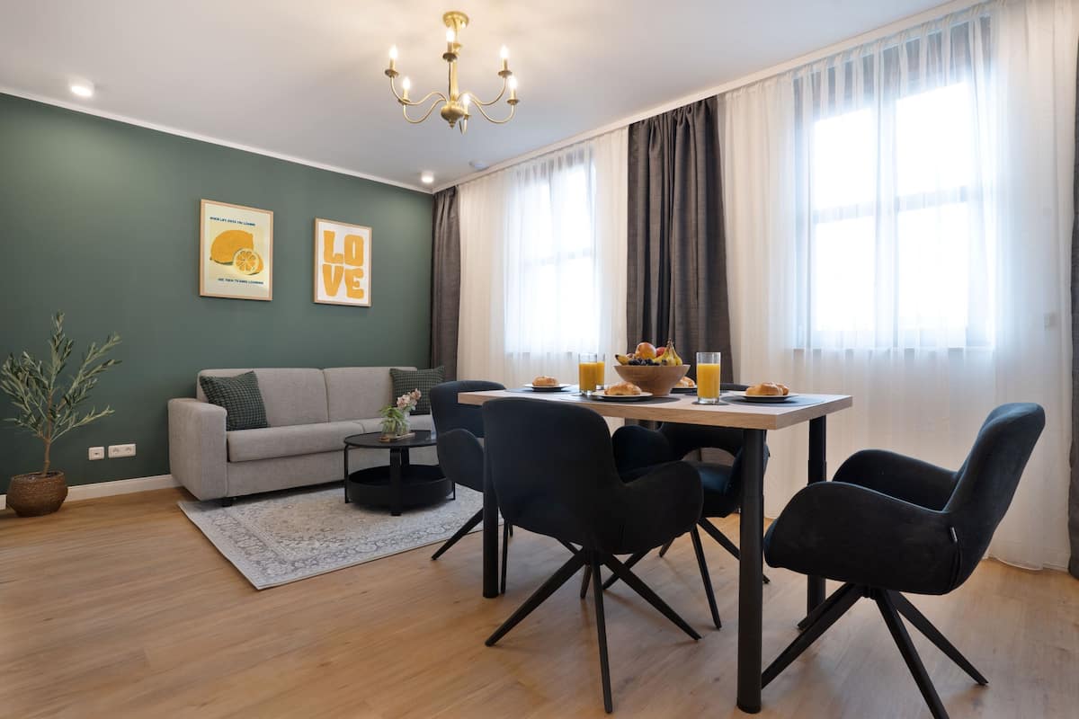 Un salon moderne avec un canapé gris, des chaises noires, une table à manger avec des fruits et des boissons, un lustre et une grande fenêtre avec des rideaux.