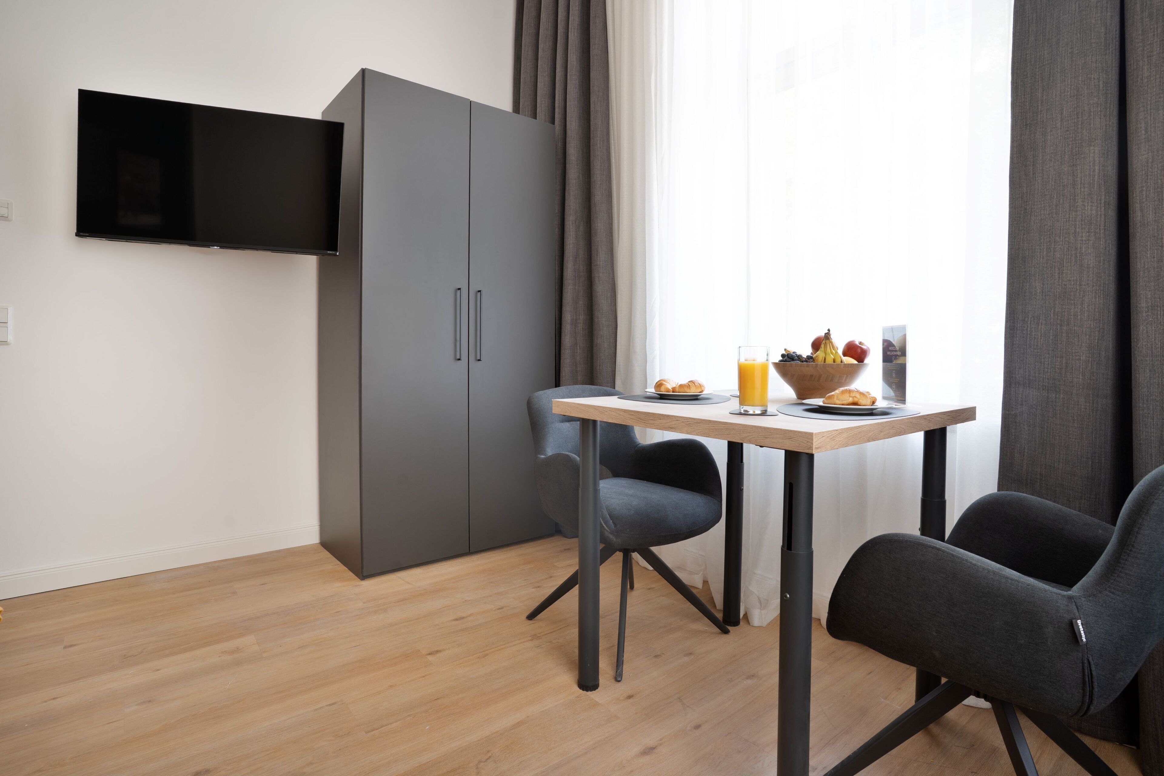 Et moderne værelse med et vægmonteret fladskærms-TV, en grå garderobe, et træspisebord med to stole og et morgenmadsmøblement med frugt og juice.