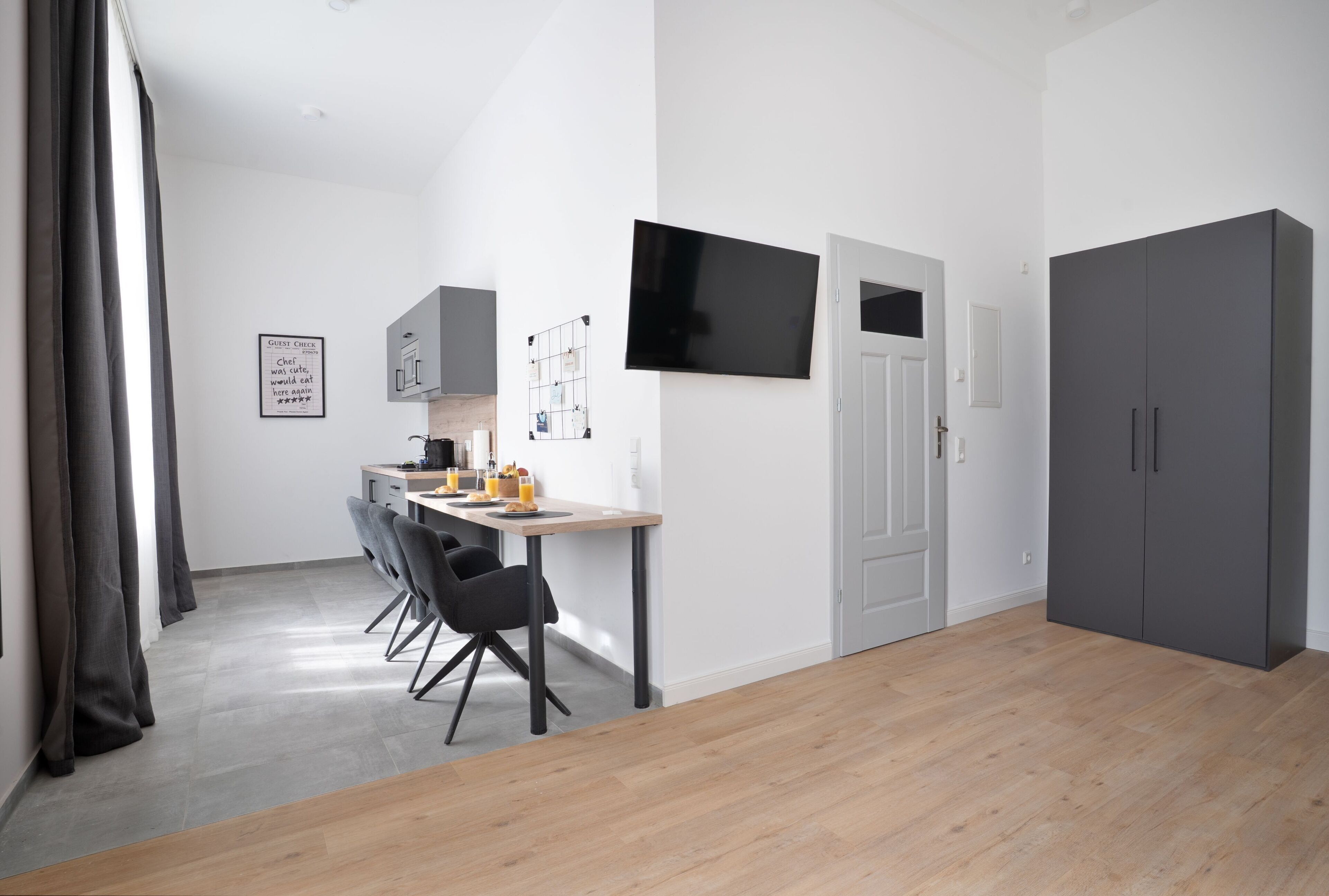 A modern, minimalist room with a dining area, a grey wardrobe, and a flat-screen TV mounted on the wall.