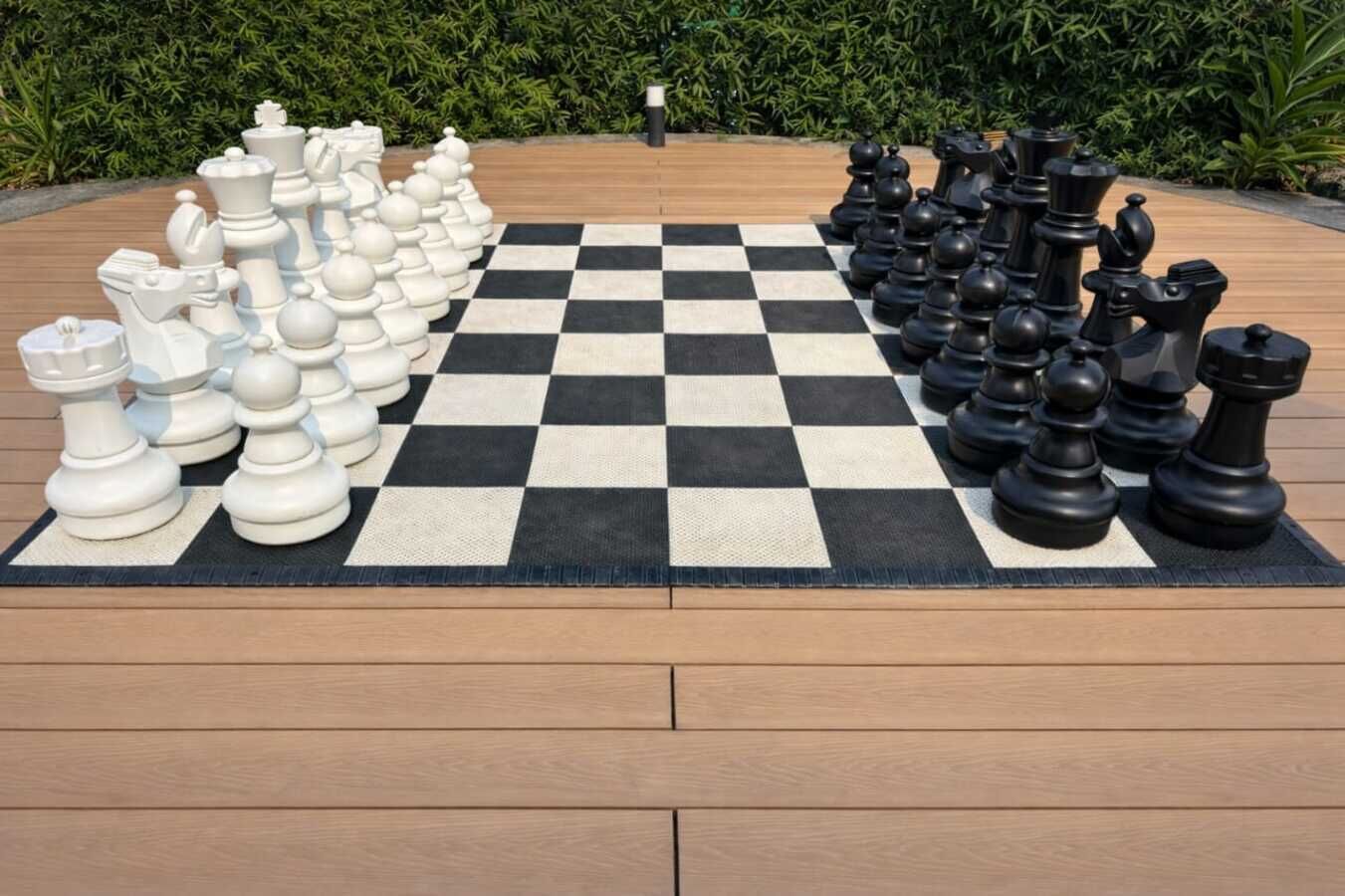A large outdoor chessboard with numerous chess pieces on a wooden deck, surrounded by greenery.