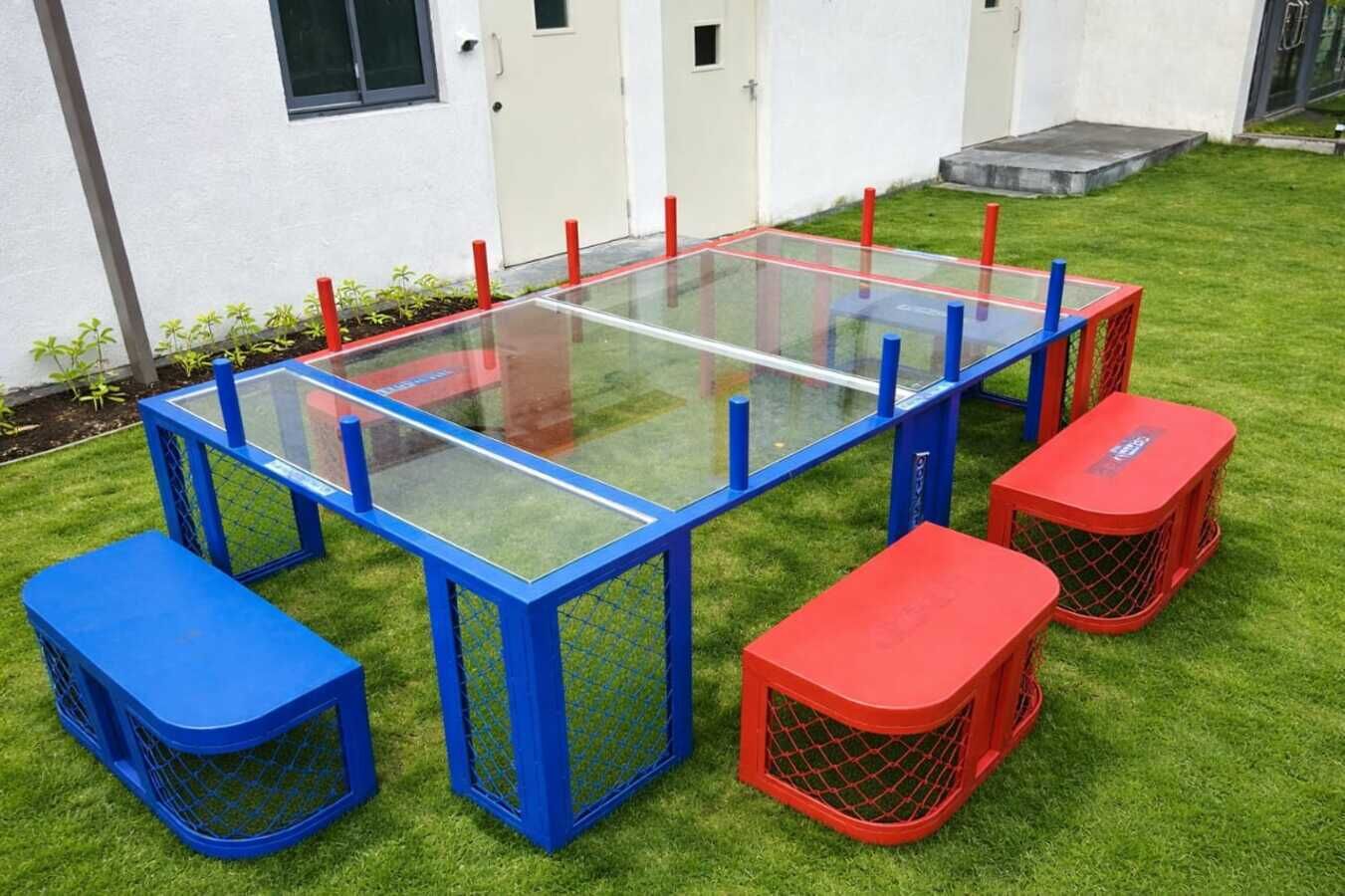 Una mesa rectangular con tapa de vidrio y patas azules y rojas, rodeada de taburetes a juego.