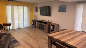 A living room with a wooden floor, a TV mounted on the wall, a fireplace, and a dining area with a table and chairs. - Driftwood Den - Walk to Folly Field Beach from Spacious 3BR/2.5BA Condo (Hilton Head Island)