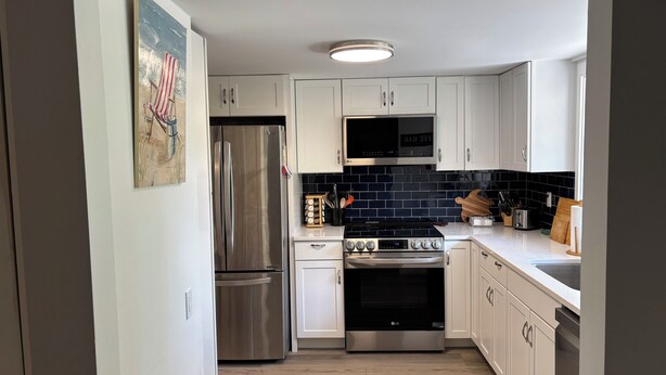 A modern kitchen with white cabinets, stainless steel appliances, a black tiled backsplash, and a wooden floor. - Driftwood Den - Walk to Folly Field Beach from Spacious 3BR/2.5BA Condo (Hilton Head Island)