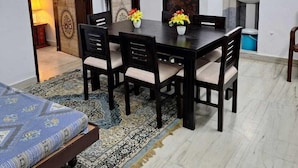 A dining area with a table and chairs, a patterned rug, and a bed with a blue cover. - Tara Home Stay (Udaipur)