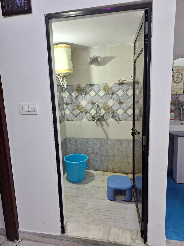 A bathroom with a shower, a blue bucket, and a small stool. - Tara Home Stay (Udaipur)