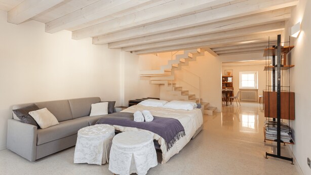 A modern bedroom with a large bed, a grey sofa, two small round tables, and a staircase leading to another room. - Large Sunny Apartment in Verona - Sleeps 8 - Easy Free Parking (Parona)