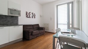 Eine moderne Küche mit weißen Schränken und einem Einbauofen, ein braunes Sofa, ein Glastisch mit zwei Stühlen sowie eine Glasschiebetür, die auf einen Balkon führt.