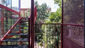 Eine rote Metalltreppe führt zu einem Balkon mit Blick auf einen Garten und Gebäude im Hintergrund.
