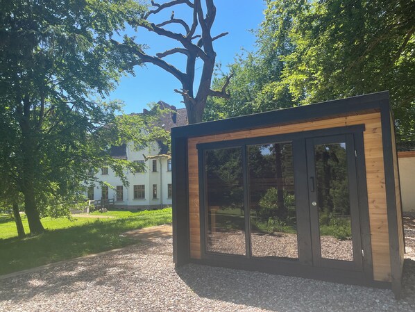 Una estructura moderna de vidrio y madera con un sendero de grava que la conduce, rodeada de árboles y un edificio blanco al fondo.