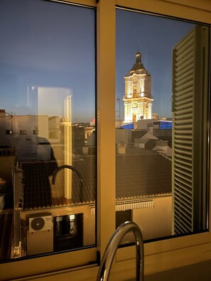 A view from a window overlooking a building with a clock tower, a tiled roof, and a window with blinds. - Madeinterranea Apartments  (Málaga)