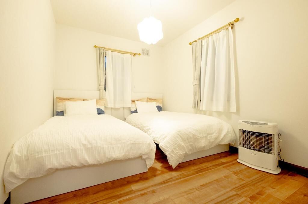 A small, well-lit bedroom with two beds, a standing heater, and a window with curtains.