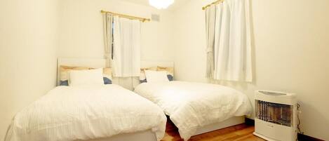 A small, well-lit bedroom with two beds, a standing heater, and a window with curtains.