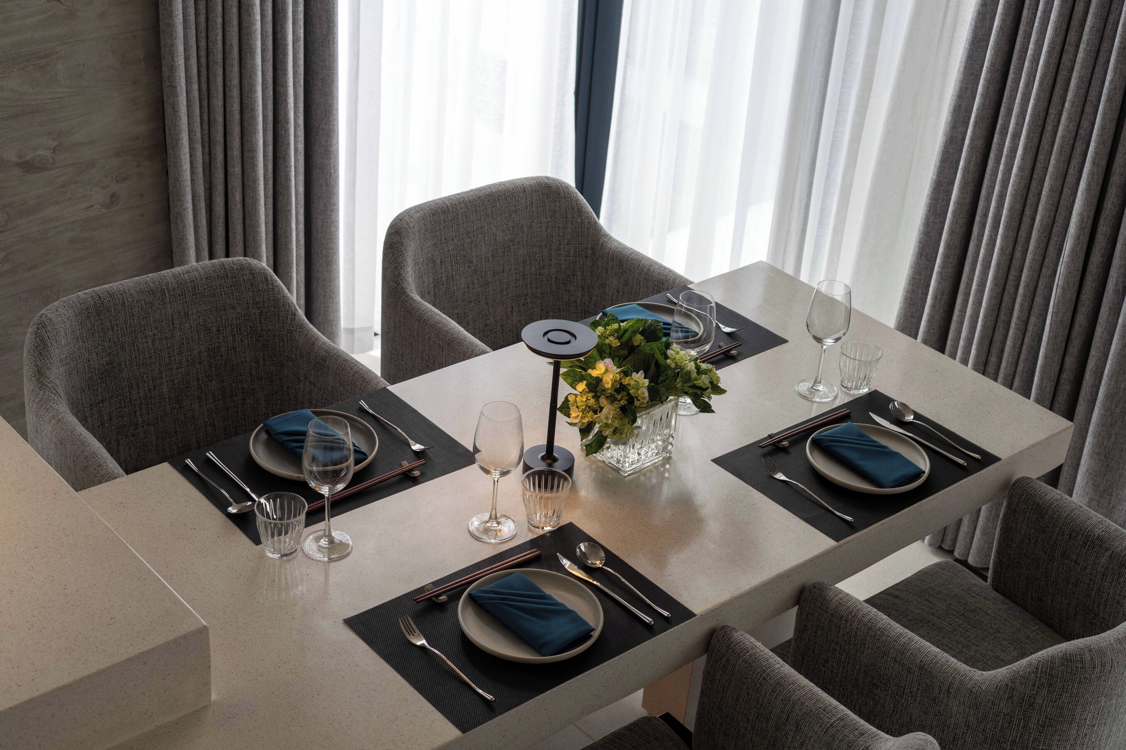 A modern dining area with a rectangular table set for four, featuring grey upholstered chairs, a vase with yellow flowers, and a minimalist decor.