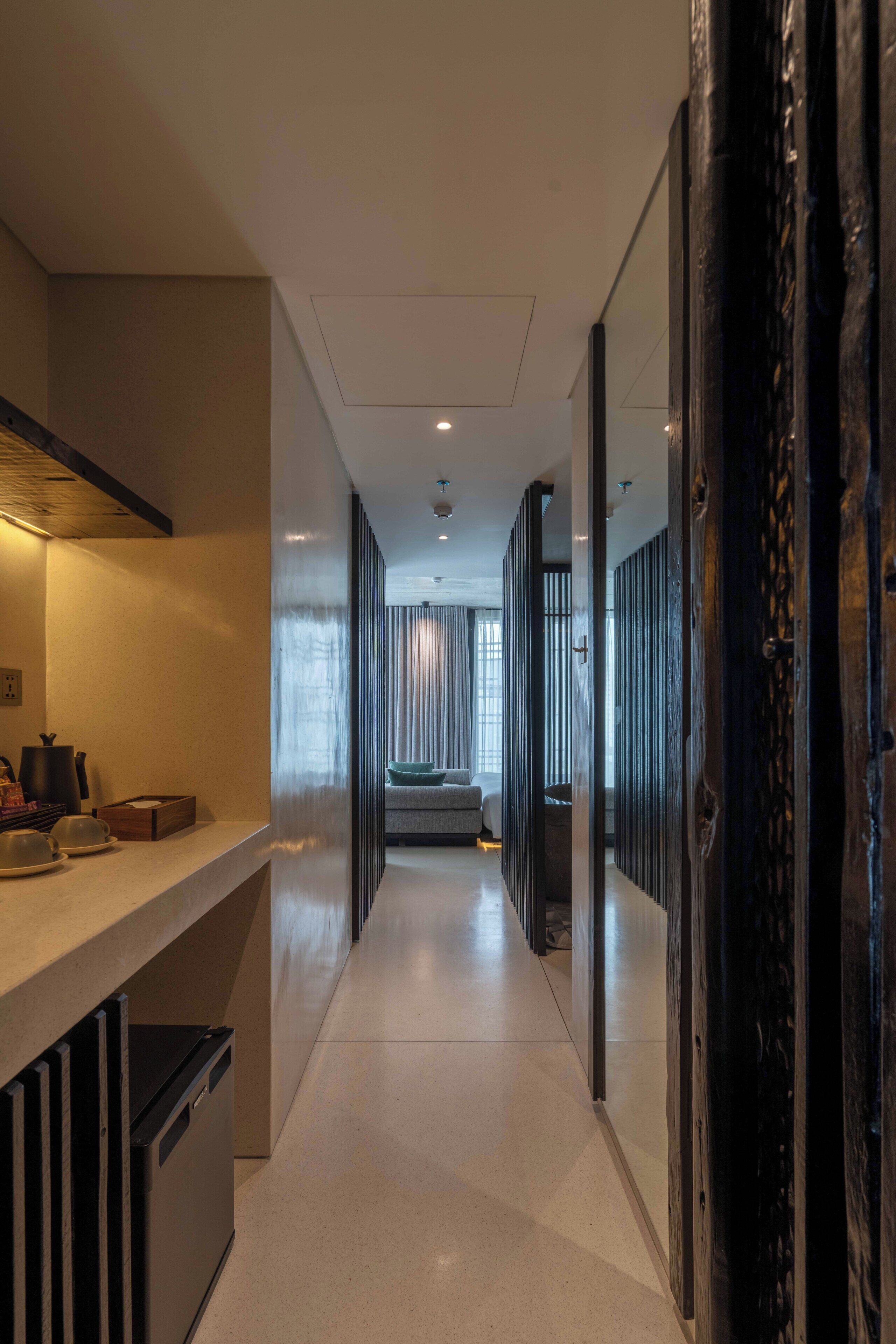 A modern hotel corridor with a kitchen area on the left, a seating area in the center, and a dark wooden door on the right.