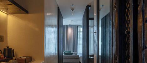 A modern hotel corridor with a kitchen area on the left, a seating area in the center, and a dark wooden door on the right.