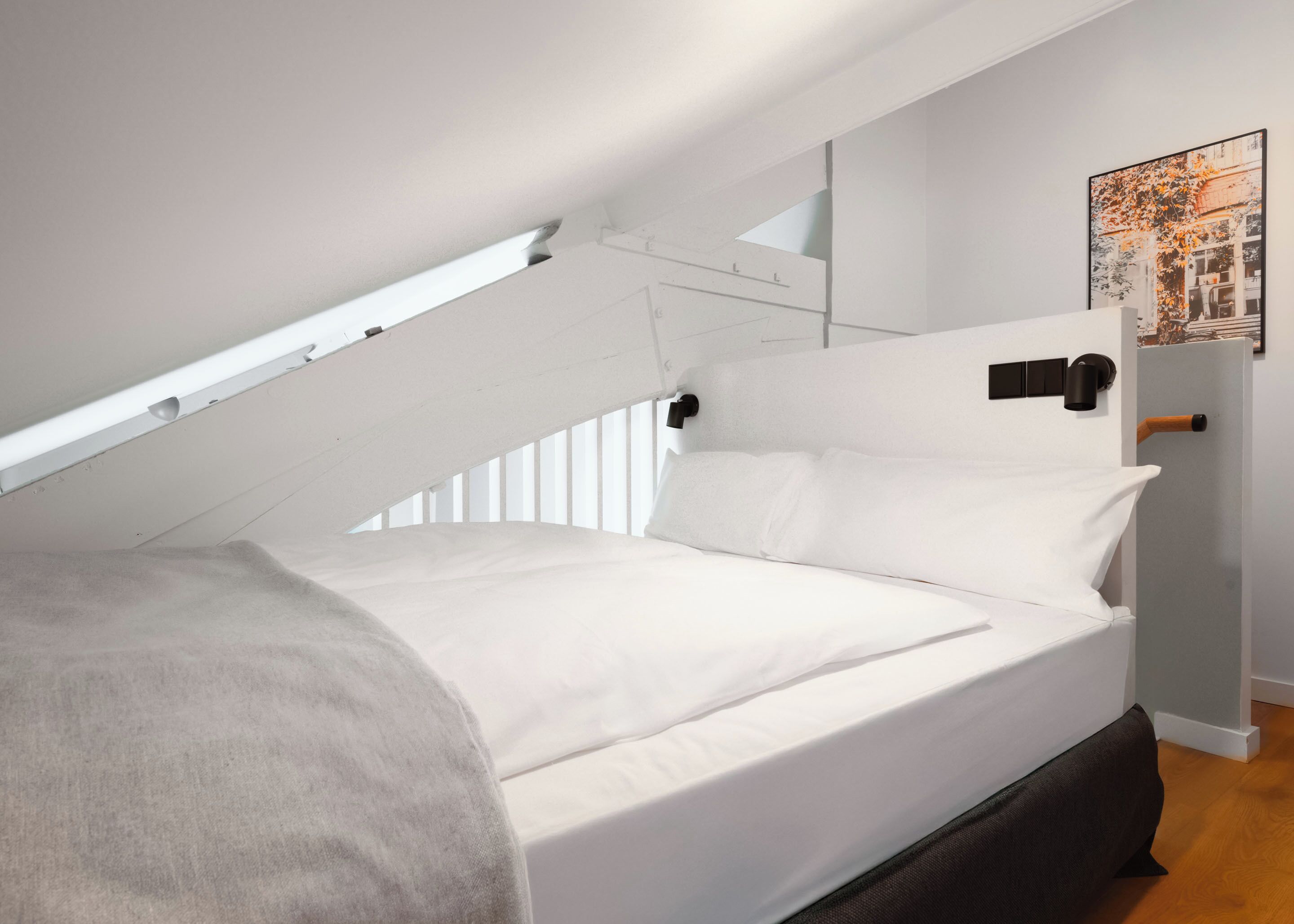 Un dormitorio con techo inclinado, una cama con ropa blanca, un plaid gris, un cabecero negro, suelo de madera e imagen enmarcada en la pared.