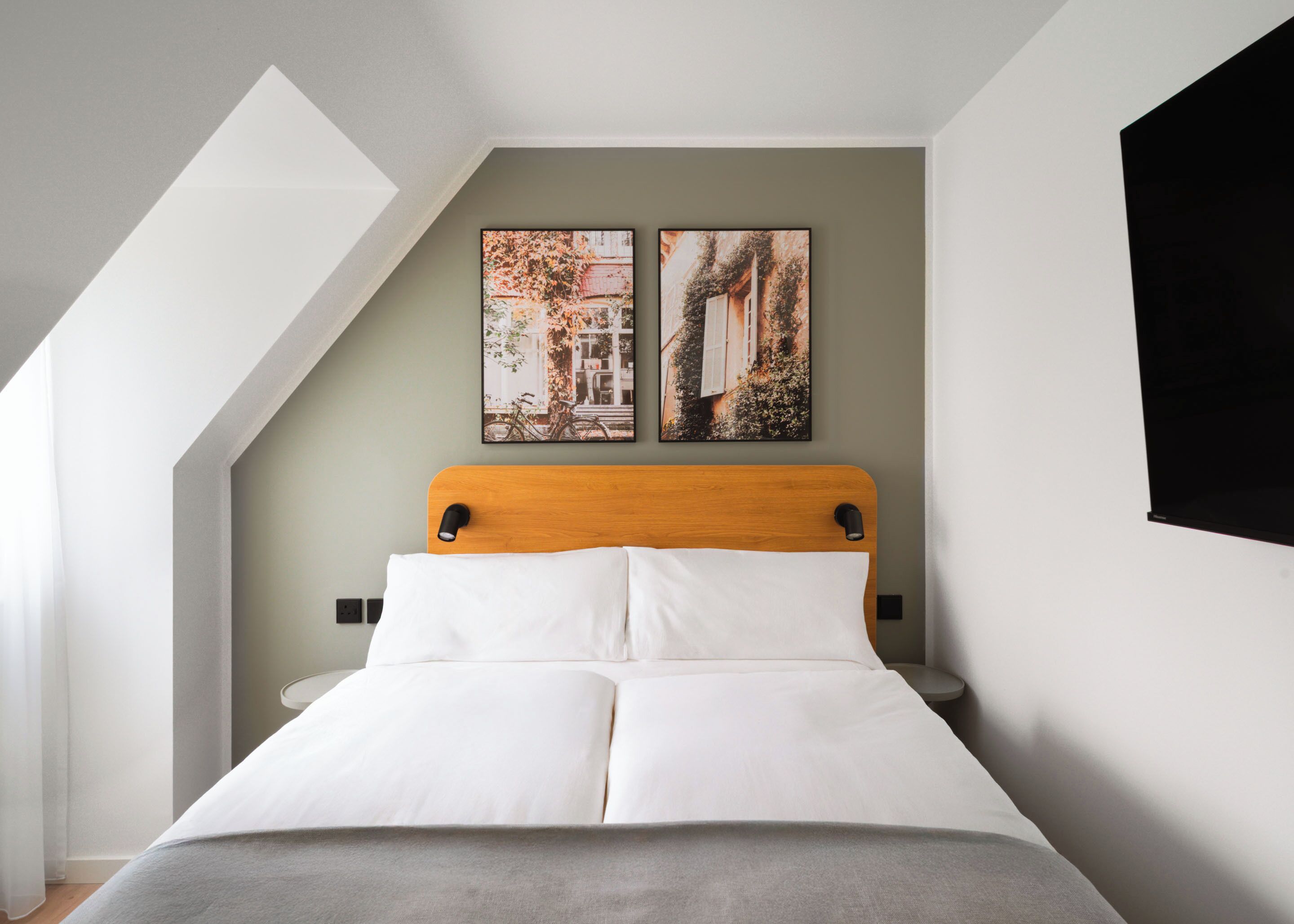 Un dormitorio con techo inclinado, cabecera de madera, cama con ropa blanca, dos cuadros colgados en la pared y un televisor negro montado en la pared.