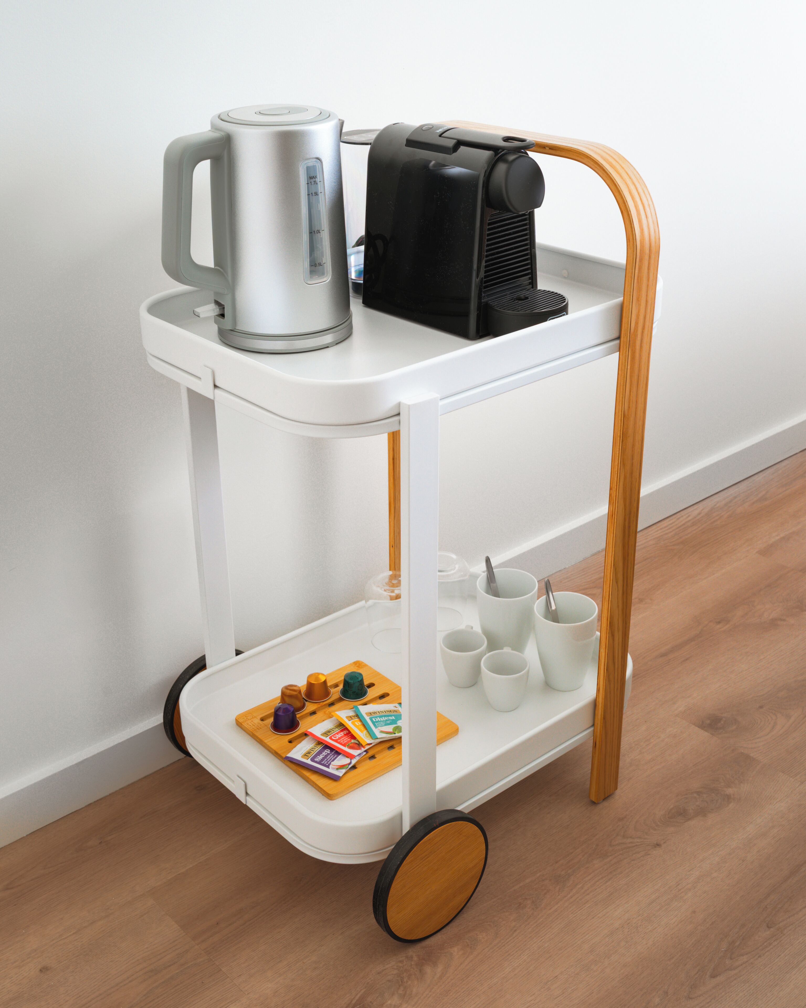 A white trolley with a wooden handle, containing a kettle, a coffee maker, and several cups.