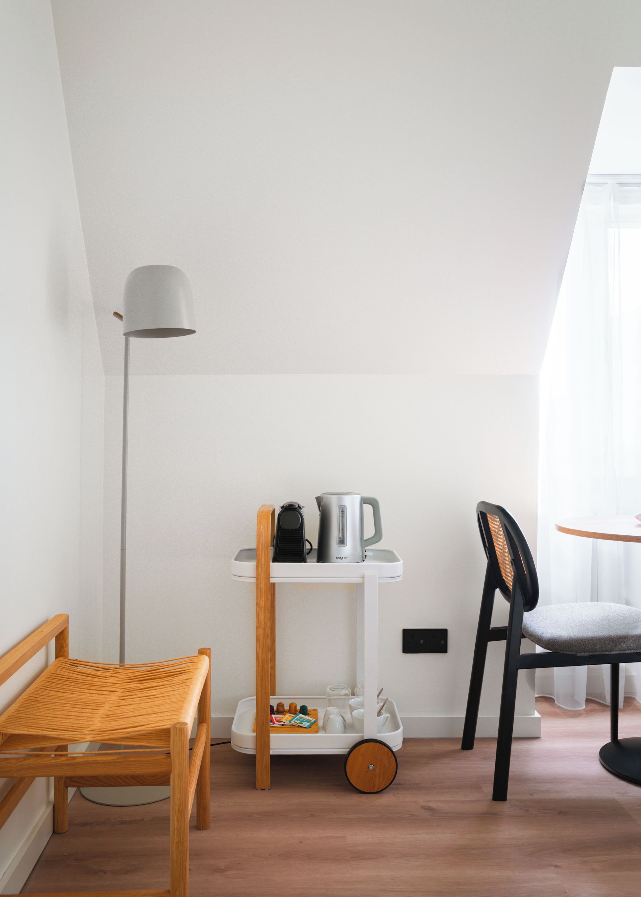 A compact room with a wooden chair, a small table, a white trolley with kitchen items, and a floor lamp.