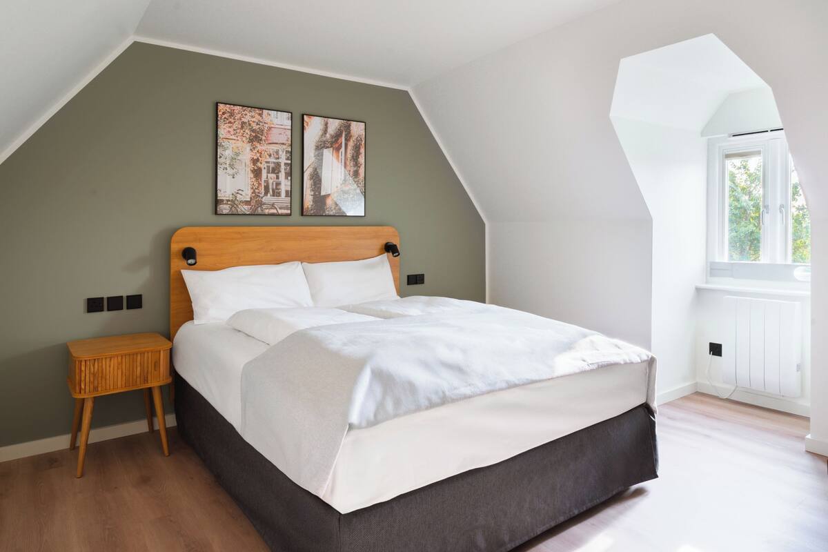 A bedroom with a sloped ceiling, a wooden headboard, a small bedside table, and two wall-mounted pictures.