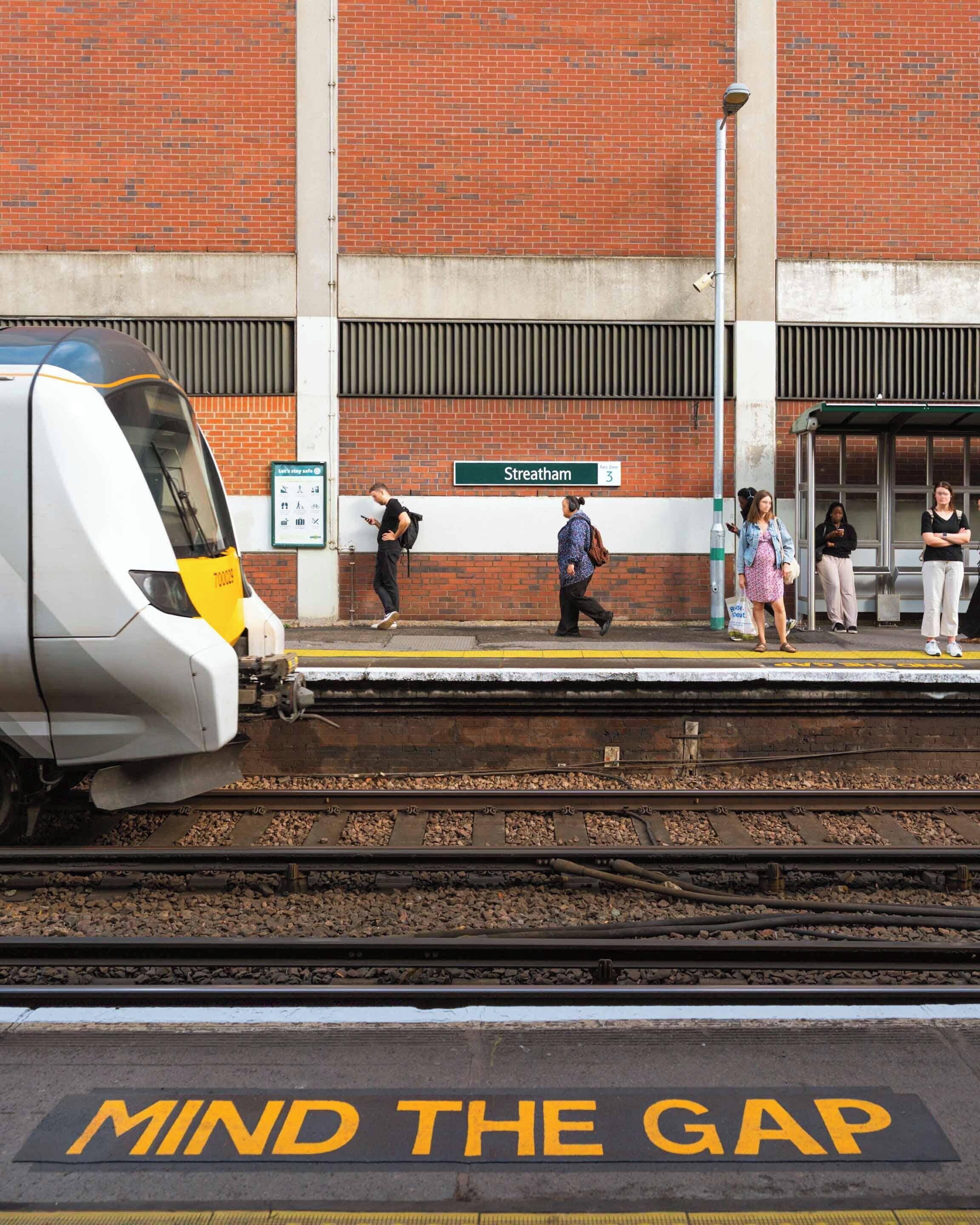 Een treinstationperron met een aankomende trein, een bord met de tekst 'Streatham' en een veiligheidswaarschuwing 'MIND THE GAP'.