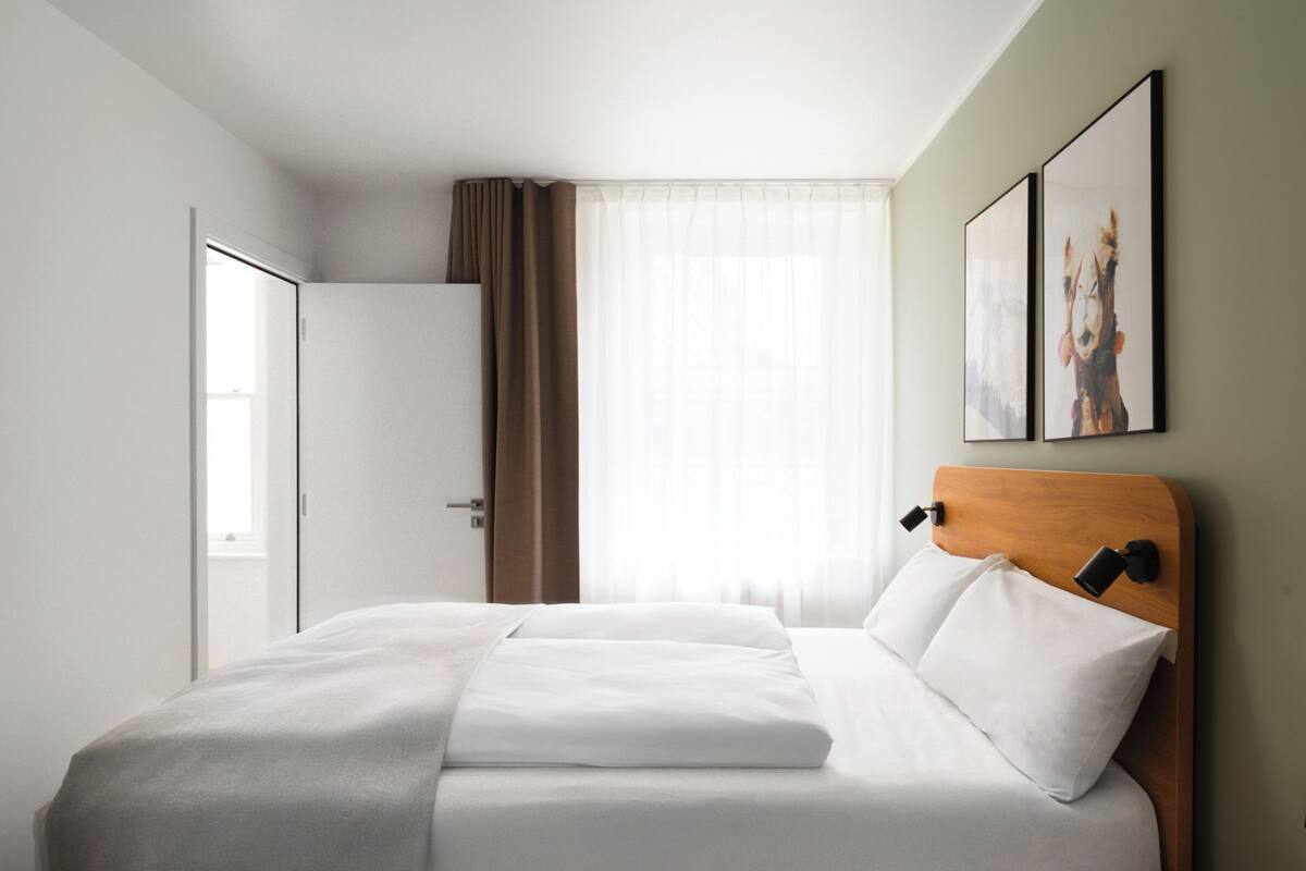 A hotel room with a neatly made bed, a wooden headboard, a bedside table, and a window with sheer curtains.