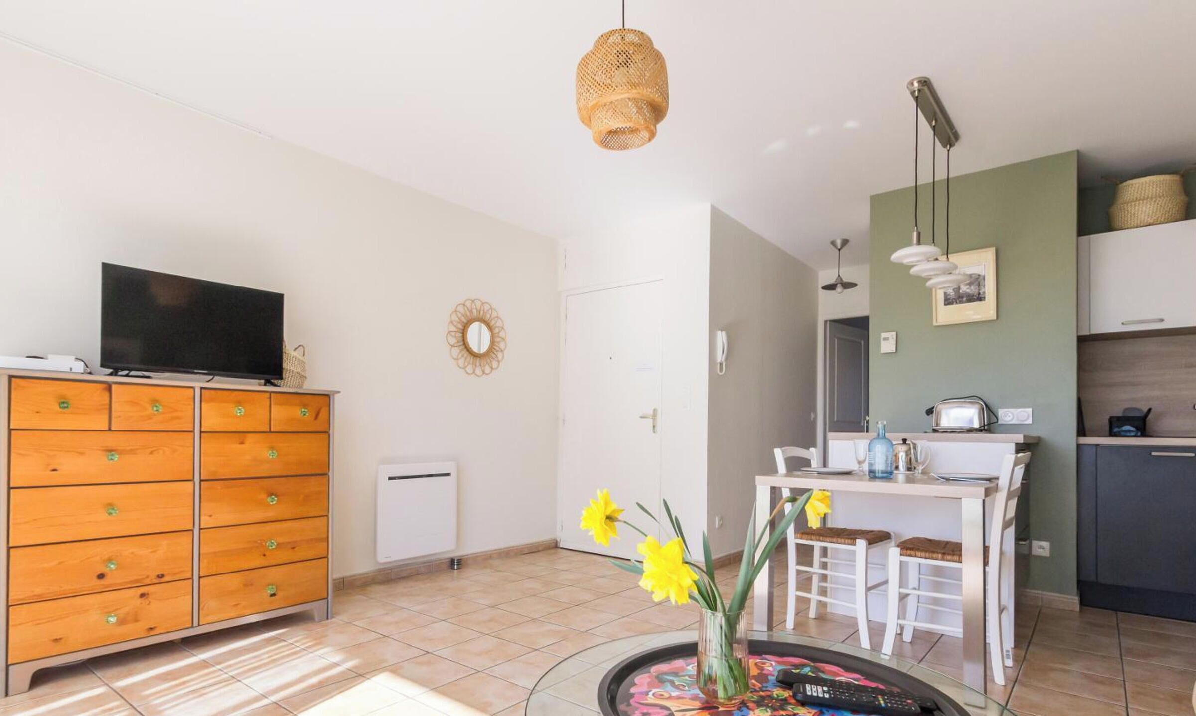 A modern living room with a TV, wooden dresser, dining table, chairs, and a kitchen area with a fridge and microwave.