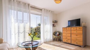 A room with a TV on a cabinet, a table with a vase and remote, and a chair with a cushion. - Charming South Garden Apartment, Pets Allowed, Parking, Barfleur (Barfleur)
