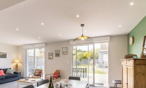 A modern living room with a sofa, dining table, and chairs, a kitchen area with a wooden cabinet, and a large sliding glass door leading to a patio.
