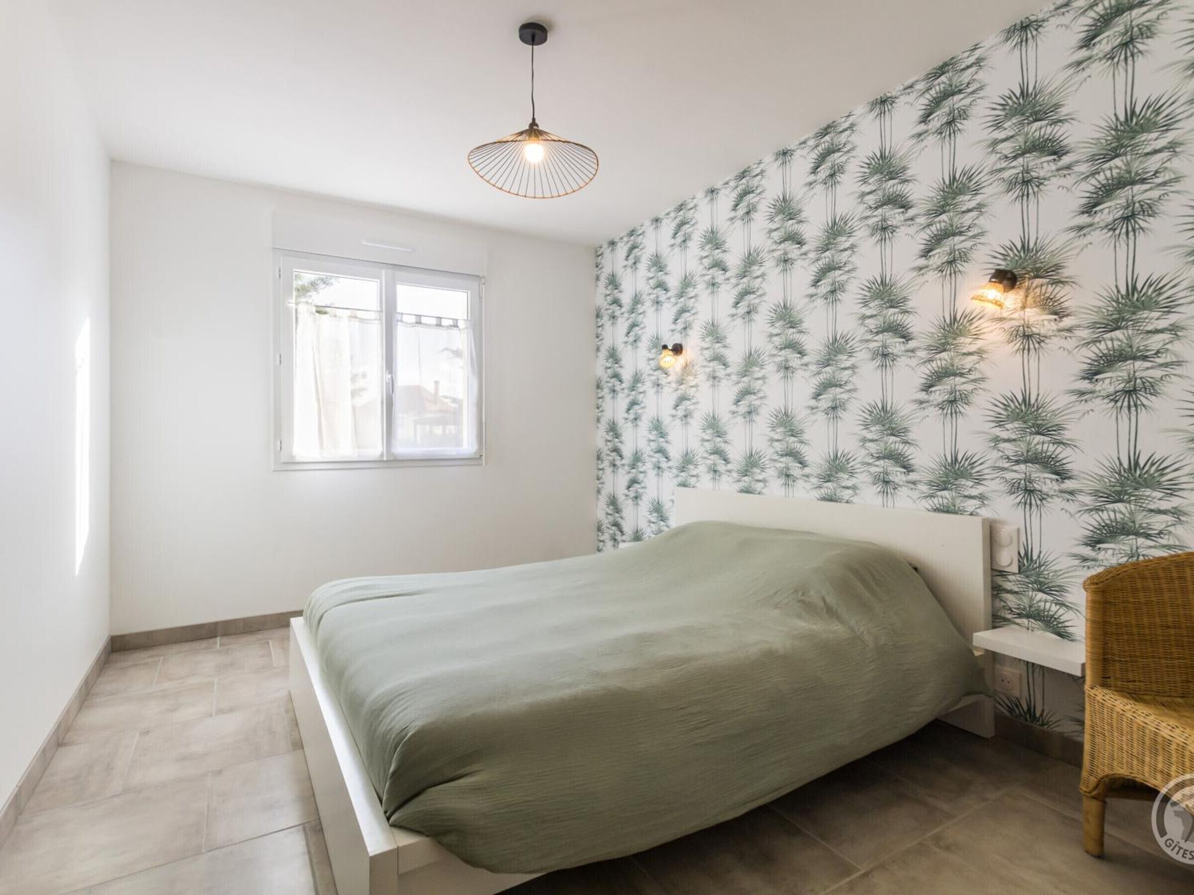 A bedroom with a bed, a window, a wall with a plant pattern, and a hanging light fixture.