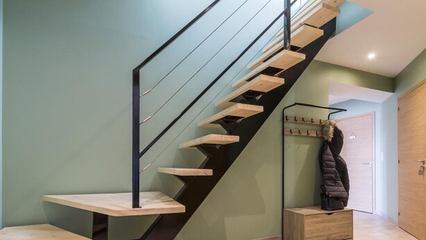 Un escalier moderne avec des marches en bois et des rampes noires, menant à une pièce où se trouvent un porte-manteau et une porte.