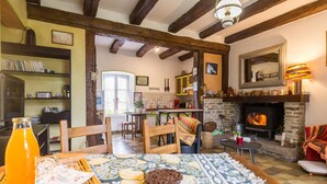 Una sala de estar acogedora con chimenea, vigas de madera, comedor y cocina visible a través de un umbral abierto.