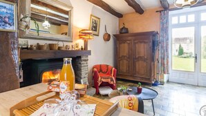 Una habitación acogedora con chimenea, vigas de madera, una mesa de comedor con vasos, un sillón rojo y vistas a un jardín a través de una puerta.