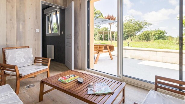 Ein modernes Zimmer mit Holzboden, einem Holztisch mit Zeitschriften, zwei Holzstühlen und einer Glastür zum Außenbereich mit Tisch und Sonnenschirm.