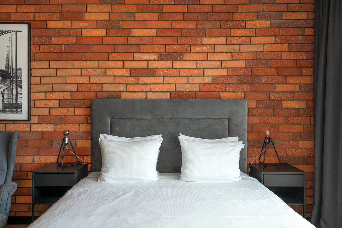 A hotel room with a brick wall, a grey upholstered headboard, two white pillows, a white bedspread, and two bedside tables with lamps.