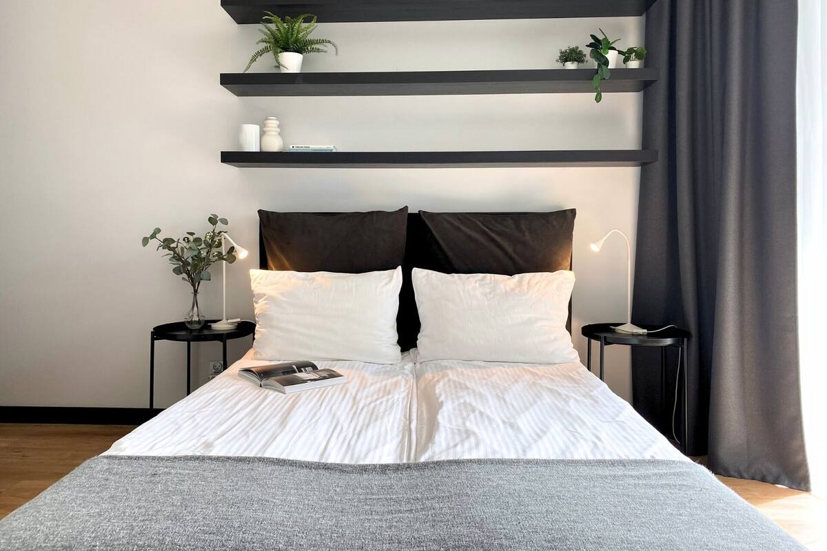 A modern bedroom with a bed, two bedside tables, a nightstand, and shelves with plants.