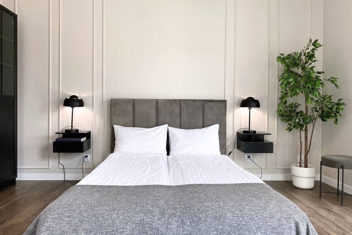 A neatly arranged bedroom with a large bed, two bedside tables with lamps, a potted plant, and a small table with a chair.