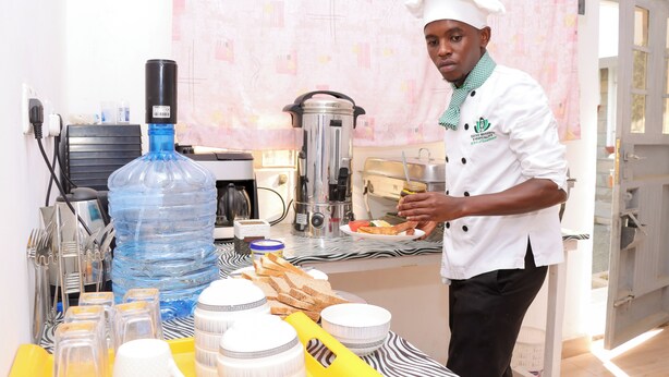 A chef in a white uniform with a green scarf, preparing food at a counter with a water dispenser, coffee machine, and various kitchen utensils. - Jazby Guest House Naivasha (Naivasha)