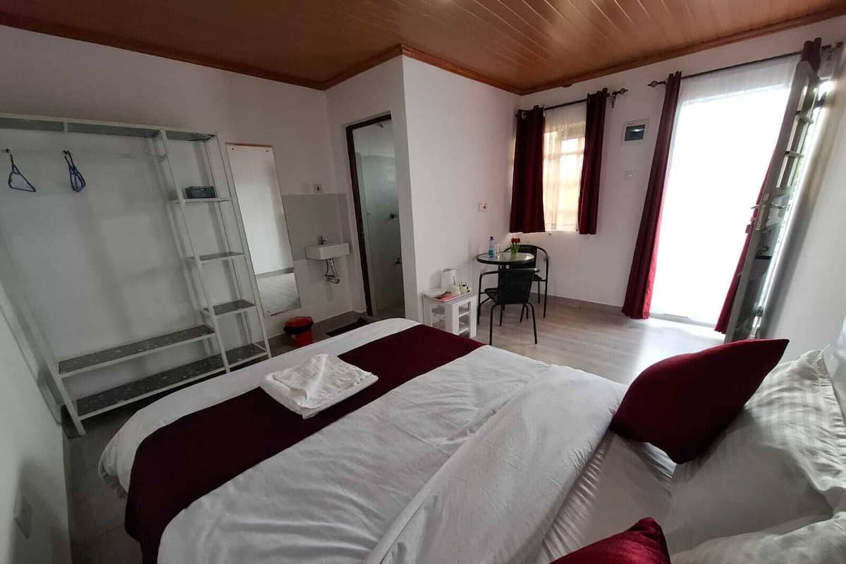 Une chambre d’hôtel avec un grand lit, un bureau, une chaise, une petite table, un miroir, une armoire et une fenêtre avec des rideaux.