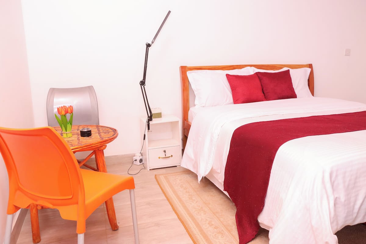 Une chambre d’hôtel avec un lit, une chaise, une petite table sur laquelle se trouve un vase de fleurs et une lampe.