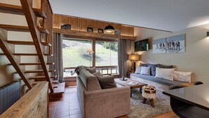 A modern living room with a sofa, coffee table, and a dining area. A wooden staircase is visible on the left, and a large window offers a view of the outdoors. - Val d'Isère - Flat for 7 ppl, 68 sqm (Val-d'Isère)