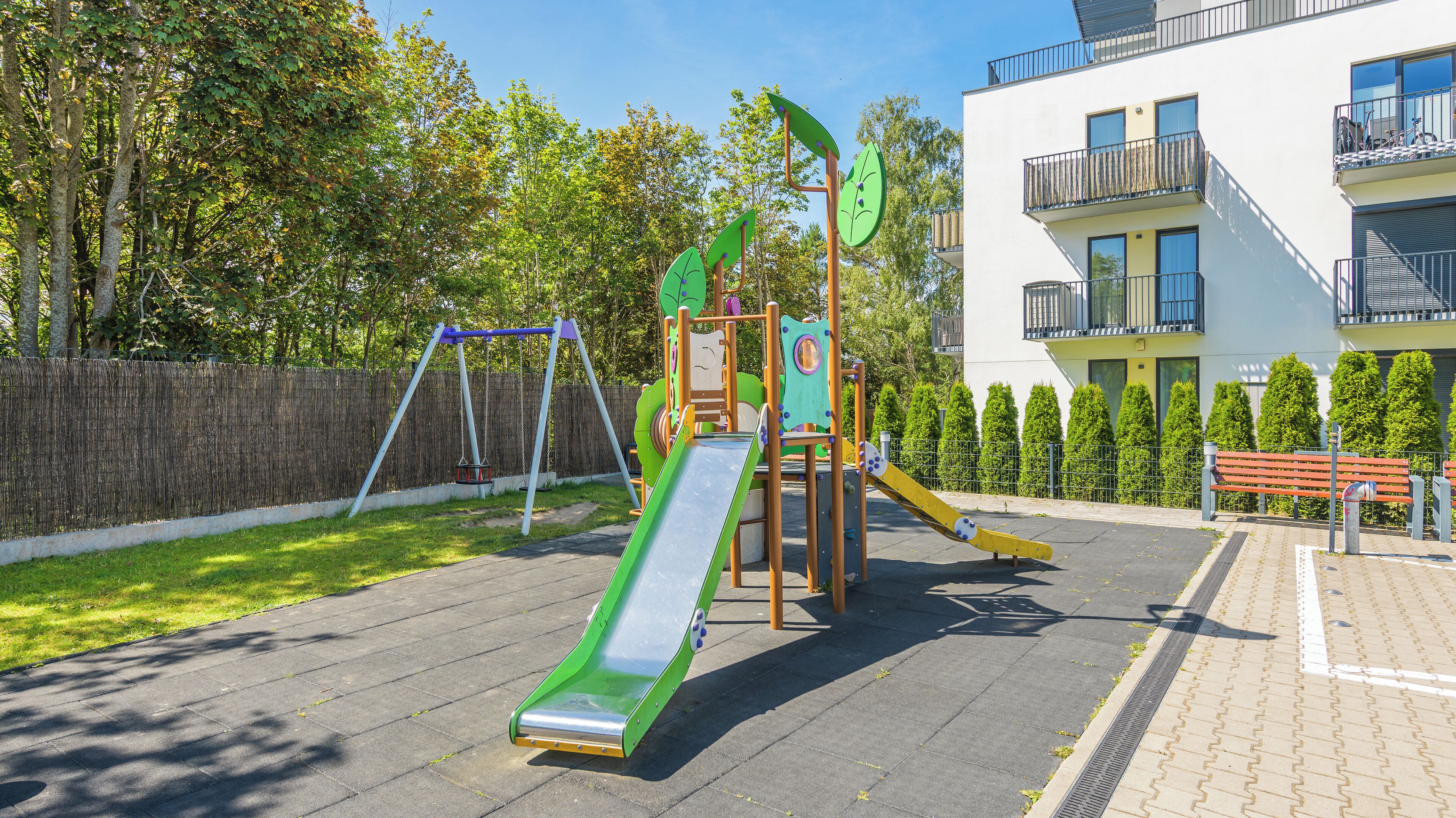Un parque infantil con tobogán y columpios, rodeado de una valla y árboles, con un edificio al fondo.