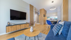 Una sala de estar moderna con un televisor de pantalla plana, armarios de madera, un sofá azul y un comedor con una cocina al fondo.