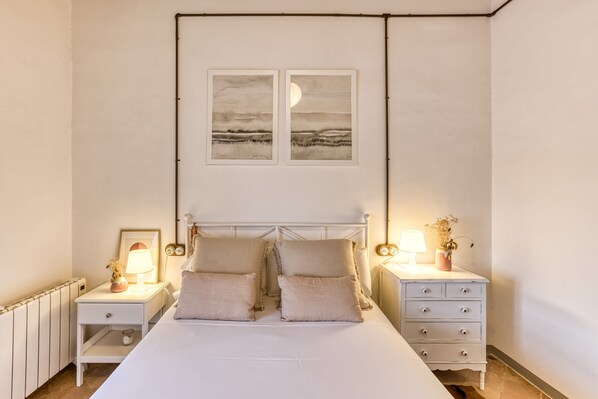 A bedroom with a white bed, two bedside tables, a nightstand, a dresser, a radiator, and two framed pictures on the wall.